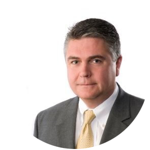 A man with short gray hair and a light complexion is wearing a gray suit jacket, white shirt, and a yellow tie. He has a neutral expression and is posing against a plain white background.