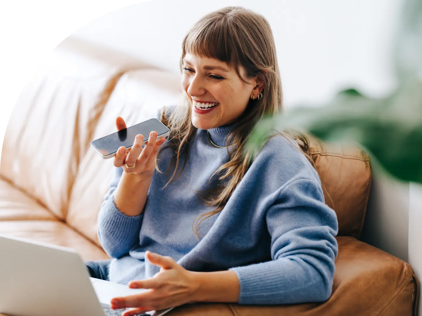 A person with long brown hair and bangs, wearing a blue sweater, is sitting on a beige leather couch. They are smiling and holding a smartphone while speaking into it about quality service, with a laptop open on their lap. The background is well-lit with a hint of greenery.